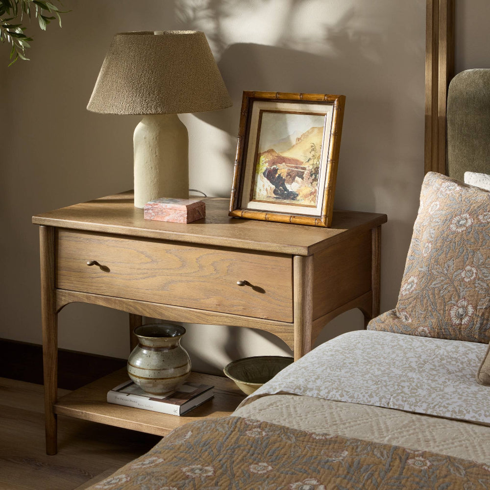 Wooden nightstand with lamp and framed picture next to a bed with patterned bedding.